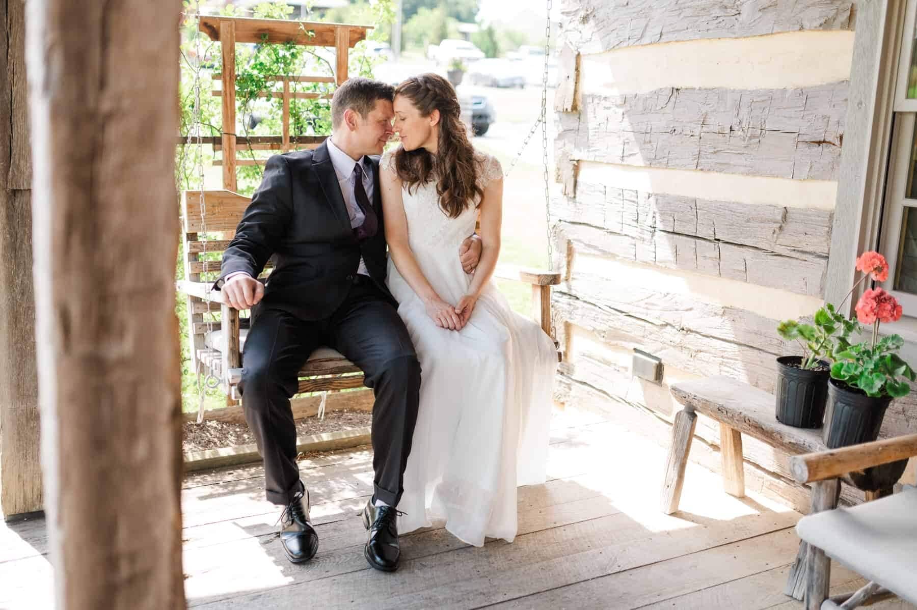 newlywed couple sit on front porch of the inn at meadow brook in swoope virginia. The two are snuggle together on the wooden porch swing of the historic, rustic cottage, nose to nose and about to share a kiss. 