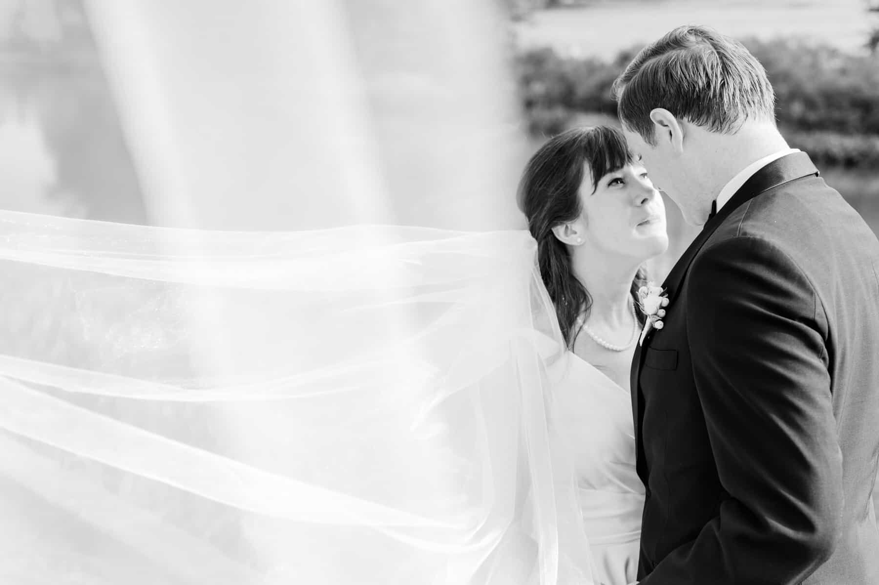 black and white image of newlywed couple under swooping cathedral wedding veil. The two are looking into each other's eyes, rather than looking at their wedding photographer.