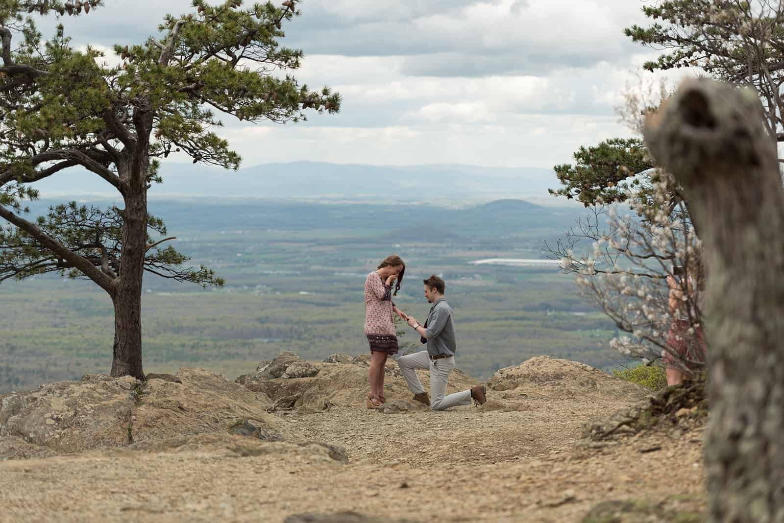 A man is down on one knee, facing his girlfriend while standing in front of the blue ridge mountain overlook on raven's roost at the skyline drive. The girlfriend is shocked and happy, with her hands on her face. He has pulled off the perfect surprise proposal plan.