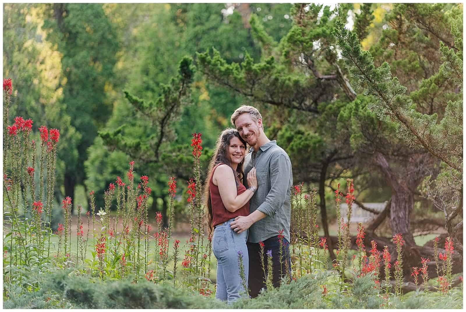 Couple cuddles up chest to chest and smiles at the camera amid red and green florals at blandy experimental farm in front royal virginia