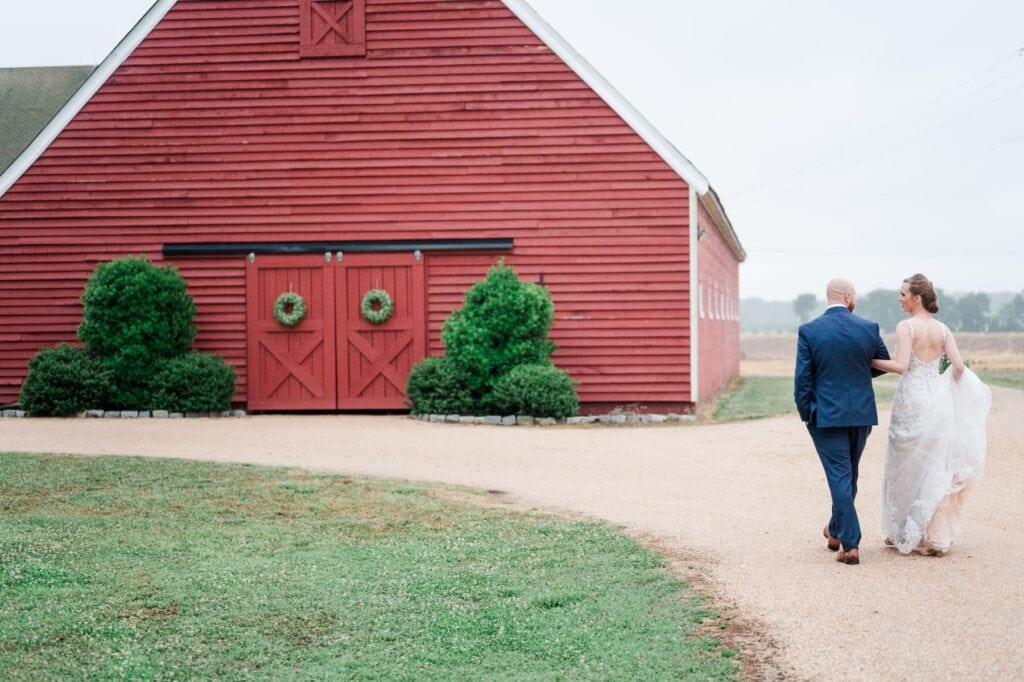 A couple walks away on a gravel path toward a large red barn, with the bride in a white dress and the groom in a dark suit.