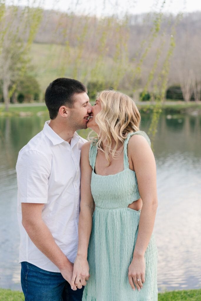 At their engagement session at Big Spring Farm, a couple stands by a serene lake, holding hands and sharing a kiss. The man is in a white shirt and jeans while the woman wears a light green dress. Behind them, trees and rolling hills complete the picturesque scene.