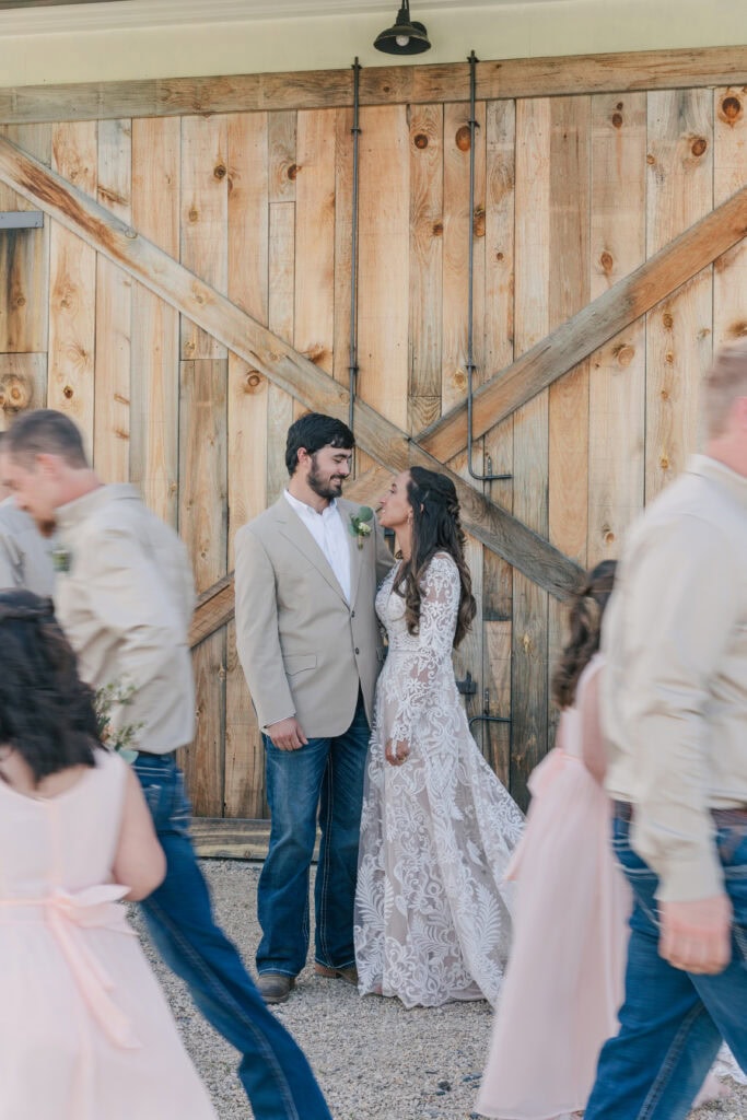 A bride and groom stand in front of a wooden barn door at Sky Ridge Farm, gazing into each other's eyes as blurred guests walk by.
