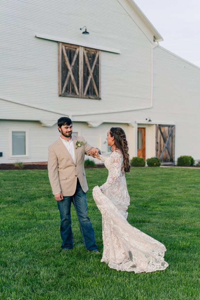 A couple dances on a grassy lawn at Sky Ridge Farm, their silhouettes framed against the barn. The man wears a beige jacket and jeans, while the woman twirls in her long white lace dress, capturing the essence of romance at this enchanting wedding venue.