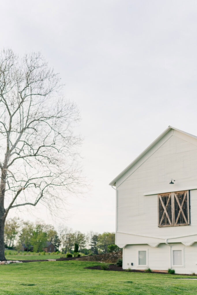 A white barn with wooden shutters on the right, a large bare tree on the left, and green grass in the foreground under a cloudy sky embodies the rustic charm of Sky Ridge Farm wedding venue.
