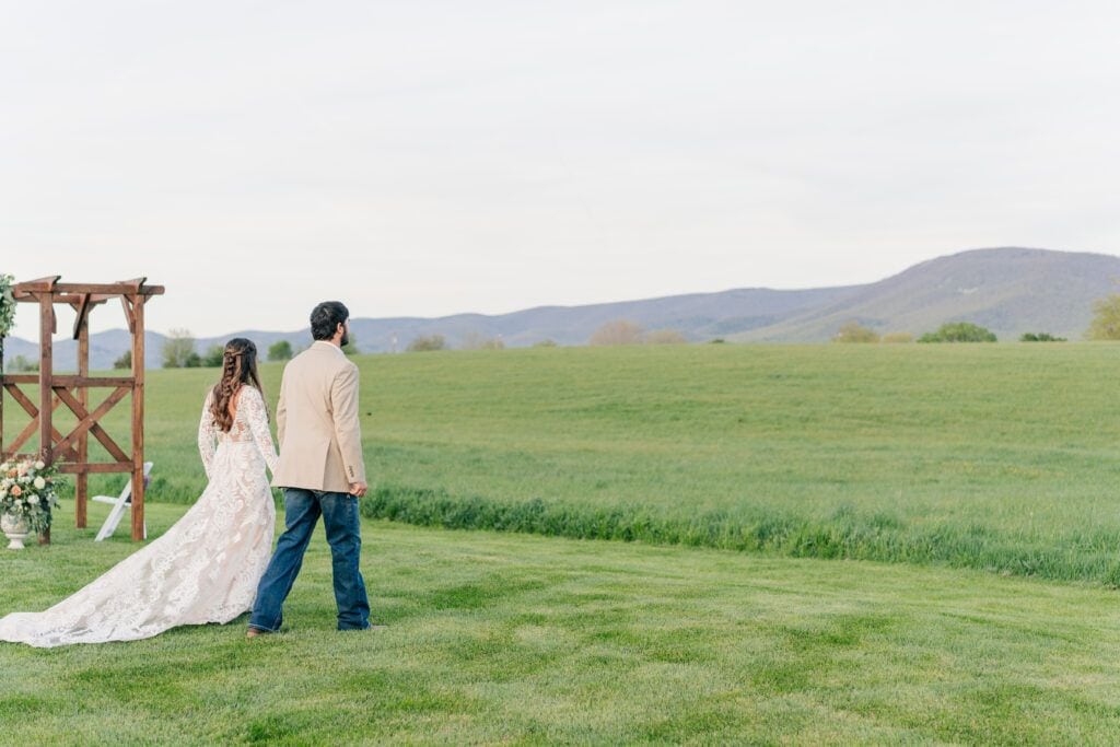 A couple, seen from behind, walks hand in hand across a grassy field towards distant mountains at Sky Ridge Farm. There is a wooden structure with flowers on the left. The sky is overcast, casting a serene ambience perfect for a wedding venue.
