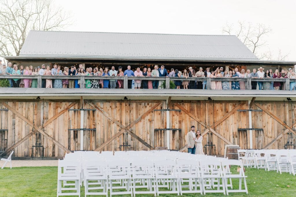 A couple stands near empty chairs in front of a wooden barn at the picturesque Sky Ridge Farm. A large group of people looks down from the barn's upper level, enjoying the view at this charming wedding venue.