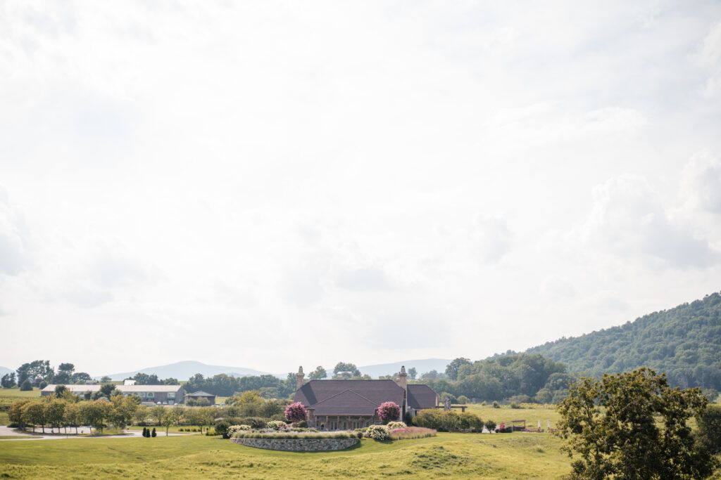 A rural landscape features a large house amidst green fields and trees, perfect as a Virginia wedding venue. It boasts distant hills and a cloudy sky, offering spectacular mountain views that make it an ideal setting for unforgettable nuptials.