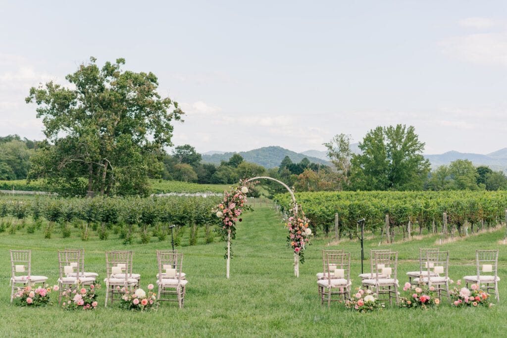 An enchanting outdoor wedding setup features a decorative arch and chairs in front of a lush vineyard under a clear sky. This picturesque Virginia wedding venue offers breathtaking mountain views, creating the perfect backdrop for your special day.