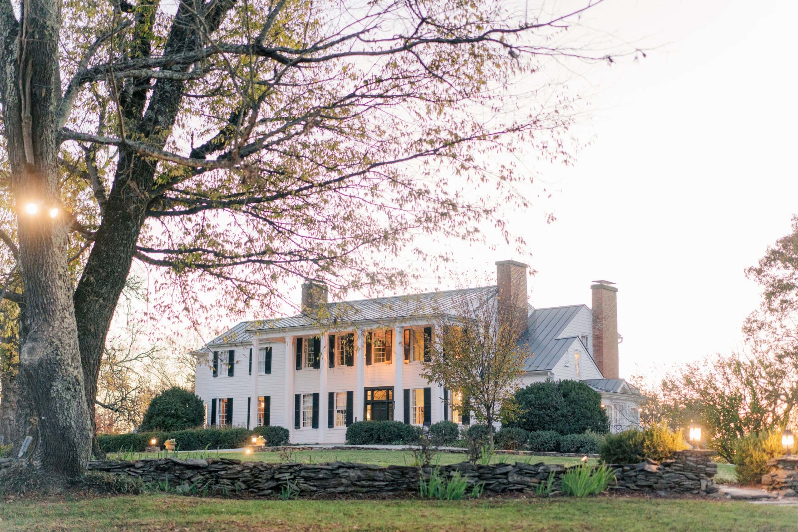 A large white colonial-style house with black shutters, perfect for a micro wedding, is surrounded by trees and a stone wall. It sits on a grassy lawn at sunset, offering an idyllic setting for intimate celebrations.