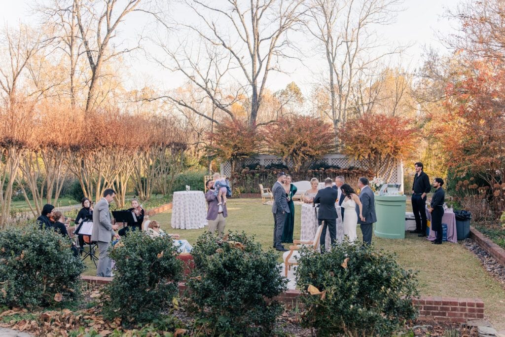 Guests gather outdoors at a Clifton Inn wedding reception, enjoying the intimate atmosphere of a micro wedding. Groups mingle around cocktail tables, surrounded by trees with autumn foliage and trimmed hedges.