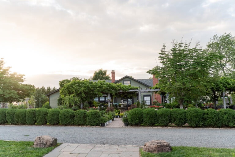 A charming house with a covered porch surrounded by lush greenery, hedges, and trees—an idyllic spot often sought after for garden wedding venues in Virginia—viewed from across a gravel driveway at sunset.