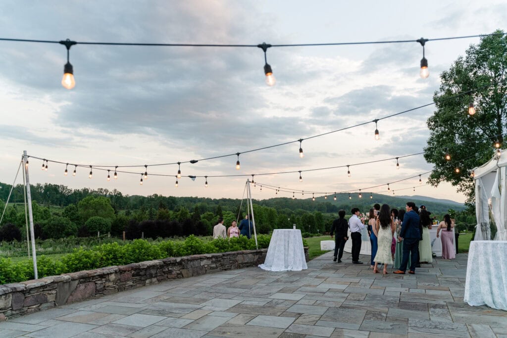 A group of people socialize outdoors under string lights at sunset, near a stone patio overlooking green trees and hills&mdash;an ideal scene for those seeking picturesque garden wedding venues in Virginia, with cocktail tables draped in white linens.
