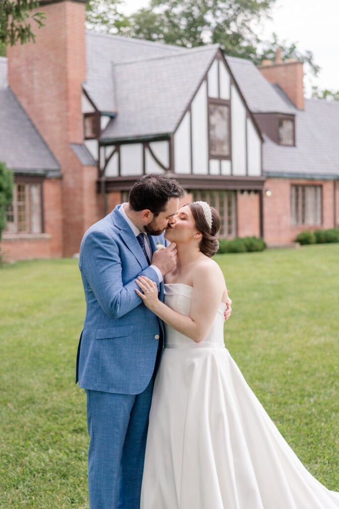 A bride in a white strapless gown and a groom in a blue suit share a kiss on the lawn of Gaie Lea, a historic wedding venue with brick and timber accents, perfect for a Staunton, Virginia wedding.