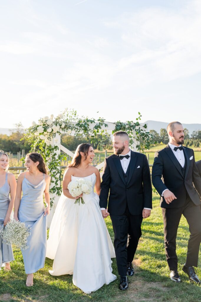 A bride and groom walk hand in hand outdoors, smiling at each other, accompanied by two bridesmaids in light blue dresses and a groomsman in a black suit, with a floral arch and green landscape in the background.