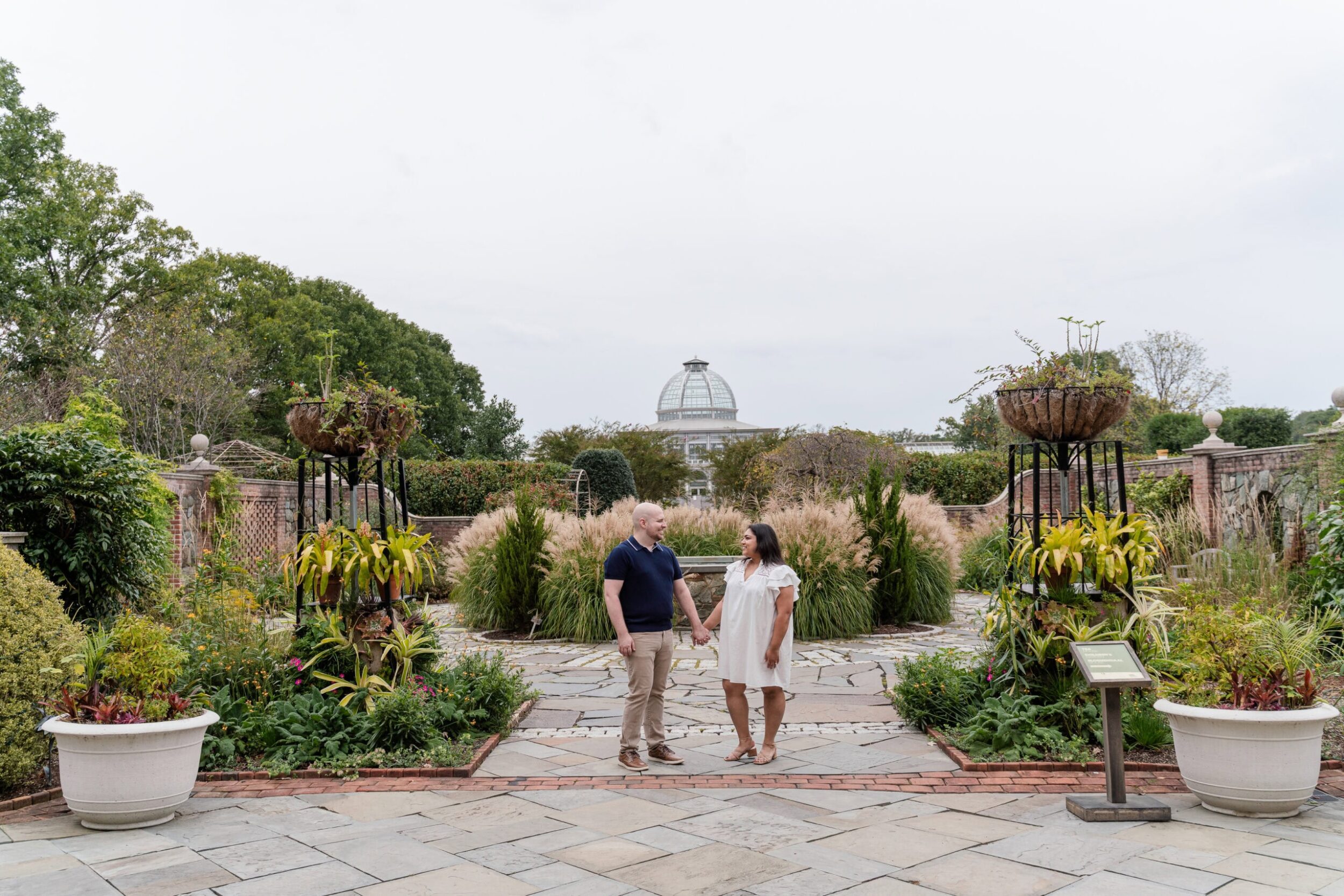 A couple stands holding hands in a lush garden with paved paths and green plants, facing each other. A domed conservatory is visible in the background under a cloudy sky.