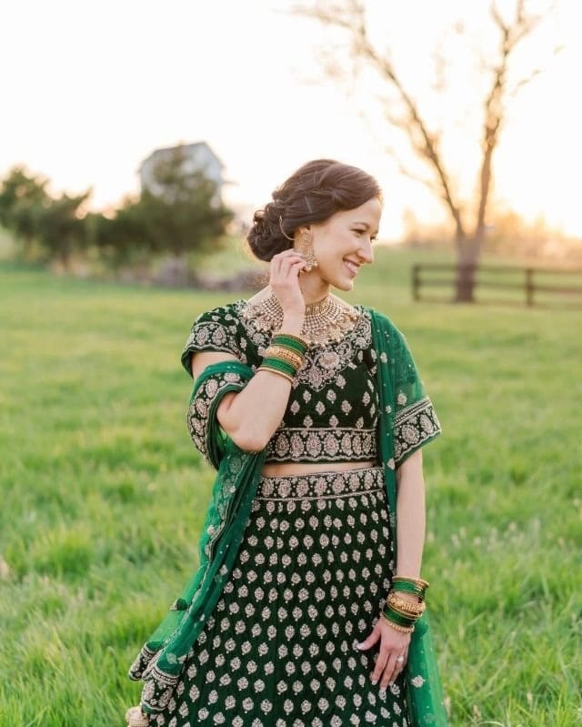 A woman in a dark green and gold traditional outfit stands in a grassy field at sunset, smiling and looking down while adjusting her earring. Trees and a fence are visible in the background.