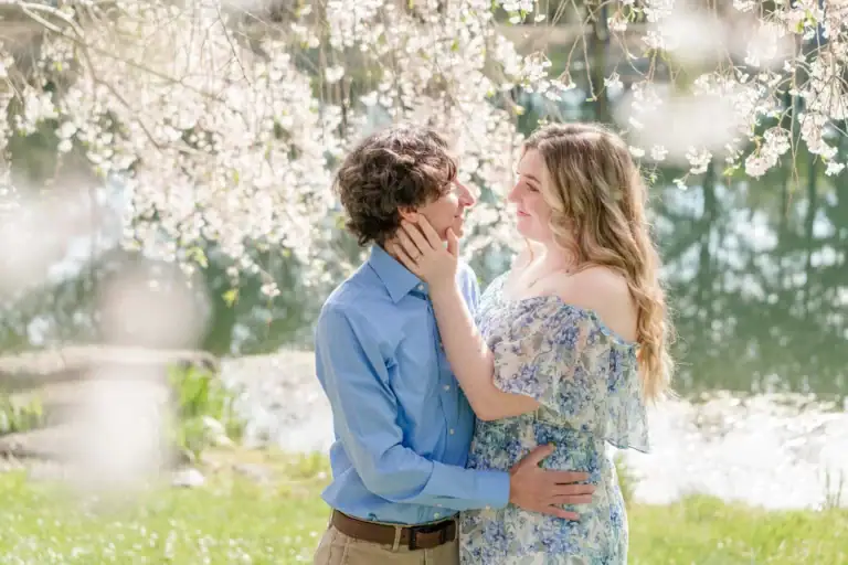 A couple stands under blooming cherry blossoms by a pond at Waterperry Farm Gardens. The woman in a floral dress gently touches the man’s face as they share a dreamy morning, surrounded by spring sunlight—a perfect Virginia engagement moment.