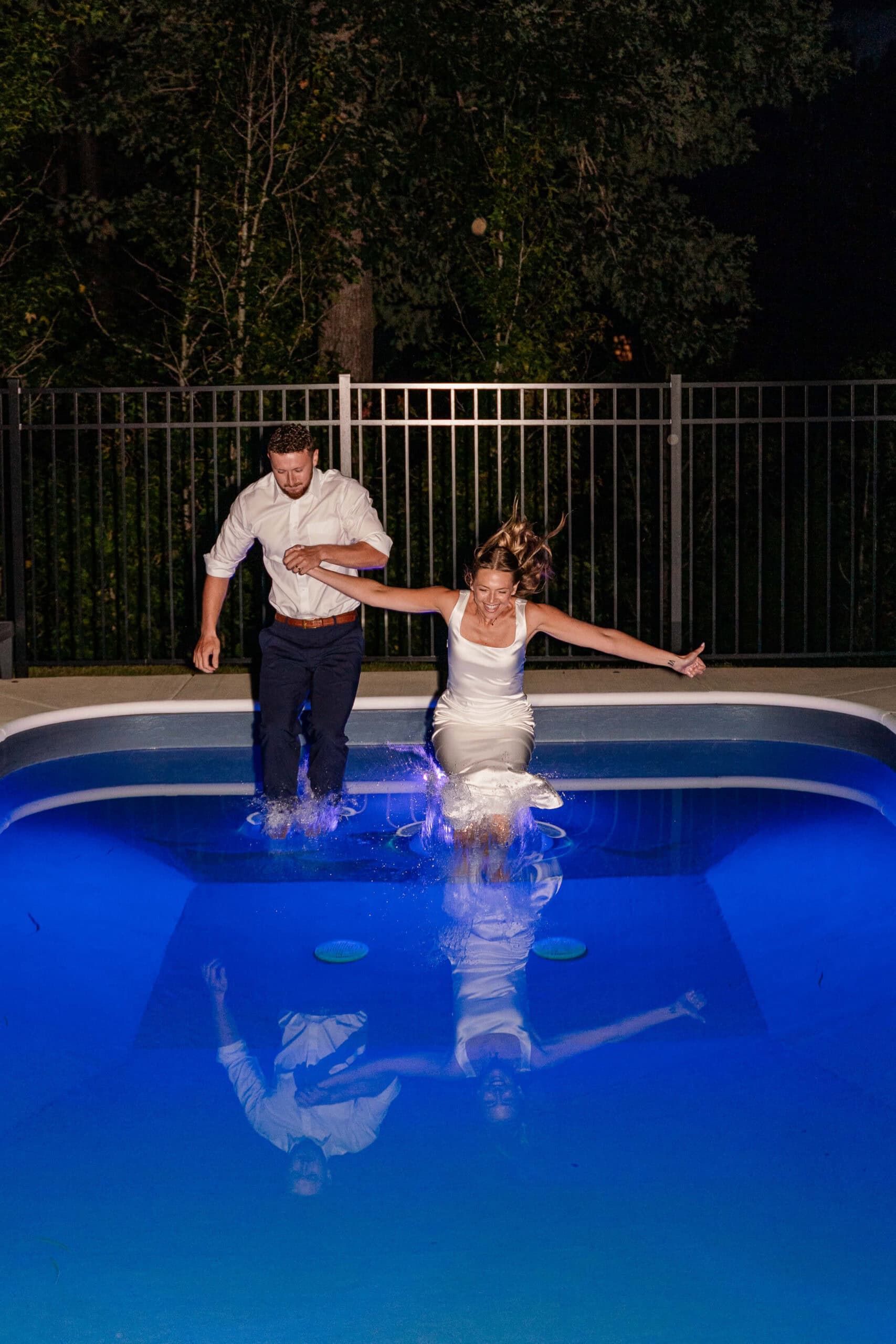 A man in a white shirt and a woman in a white dress are jumping into a brightly lit pool at night, creating a splash. There is a black fence and trees in the background.
