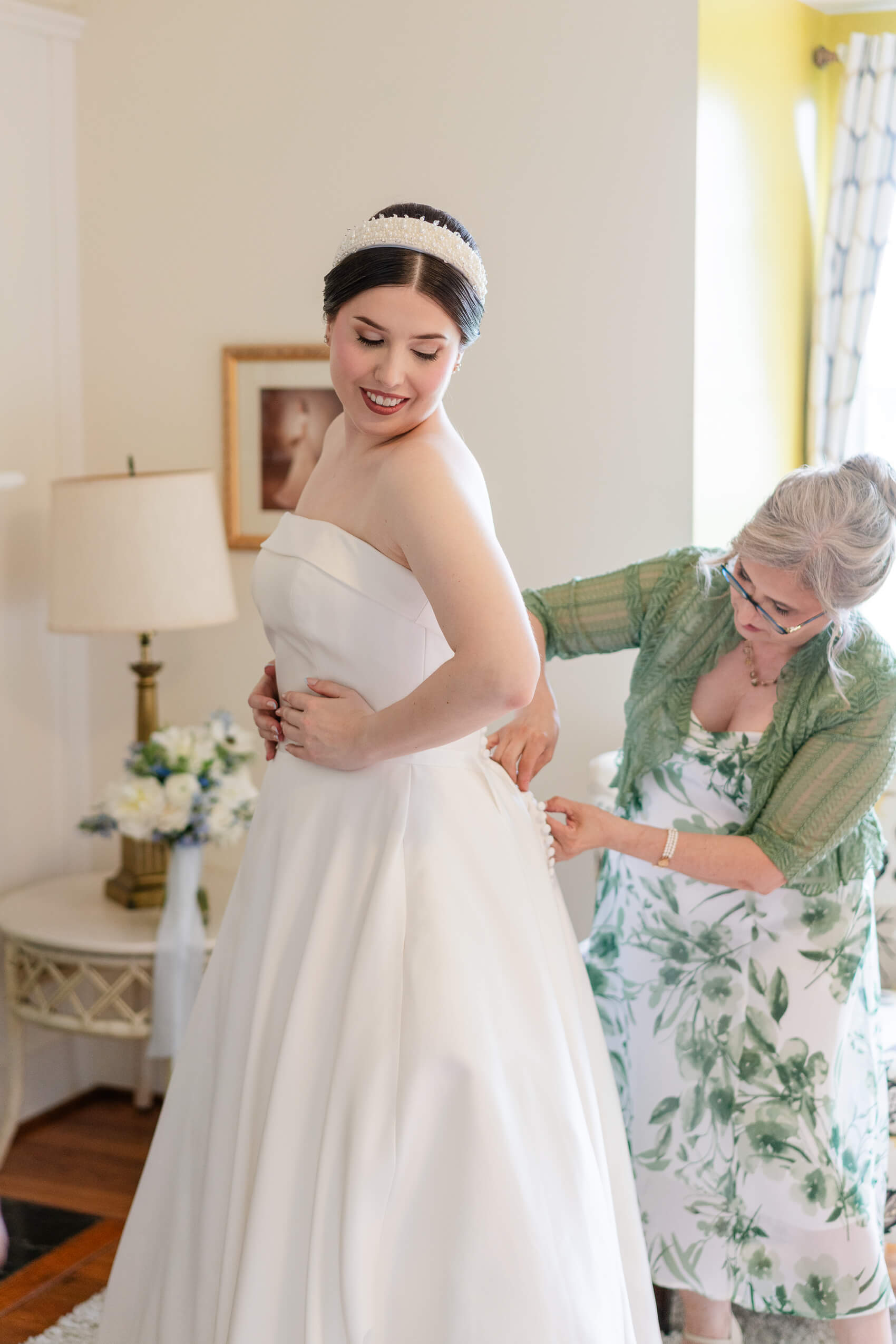 A bride in a white strapless gown smiles as an older woman in a green floral dress helps fasten the back of her dress in a softly lit room. Flowers and a lamp are visible in the background.