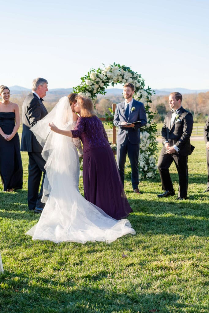 A bride in a white gown is embraced by a woman in a purple dress during an outdoor wedding ceremony. An officiant and wedding party stand nearby, with a floral arch and mountains in the background.