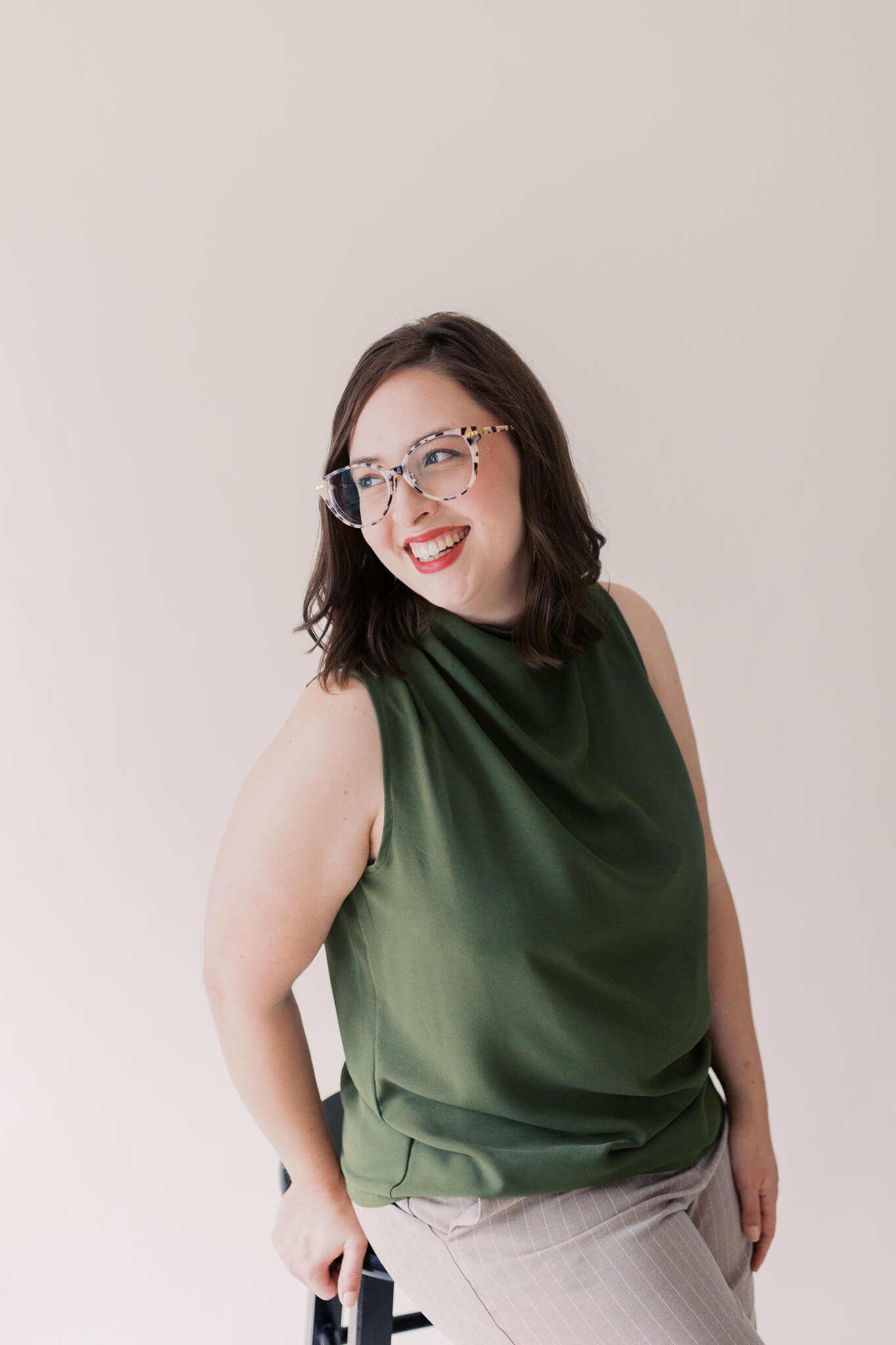 A woman with shoulder-length brown hair, wearing glasses, a sleeveless olive green top, and light-colored pants, sits smiling and looking to the side against a plain, light background.