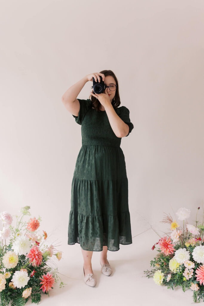 A woman in a dark green dress stands on a light floor, holding a camera up to her face. She is surrounded by colorful flower arrangements on either side. The background is plain and light-colored.