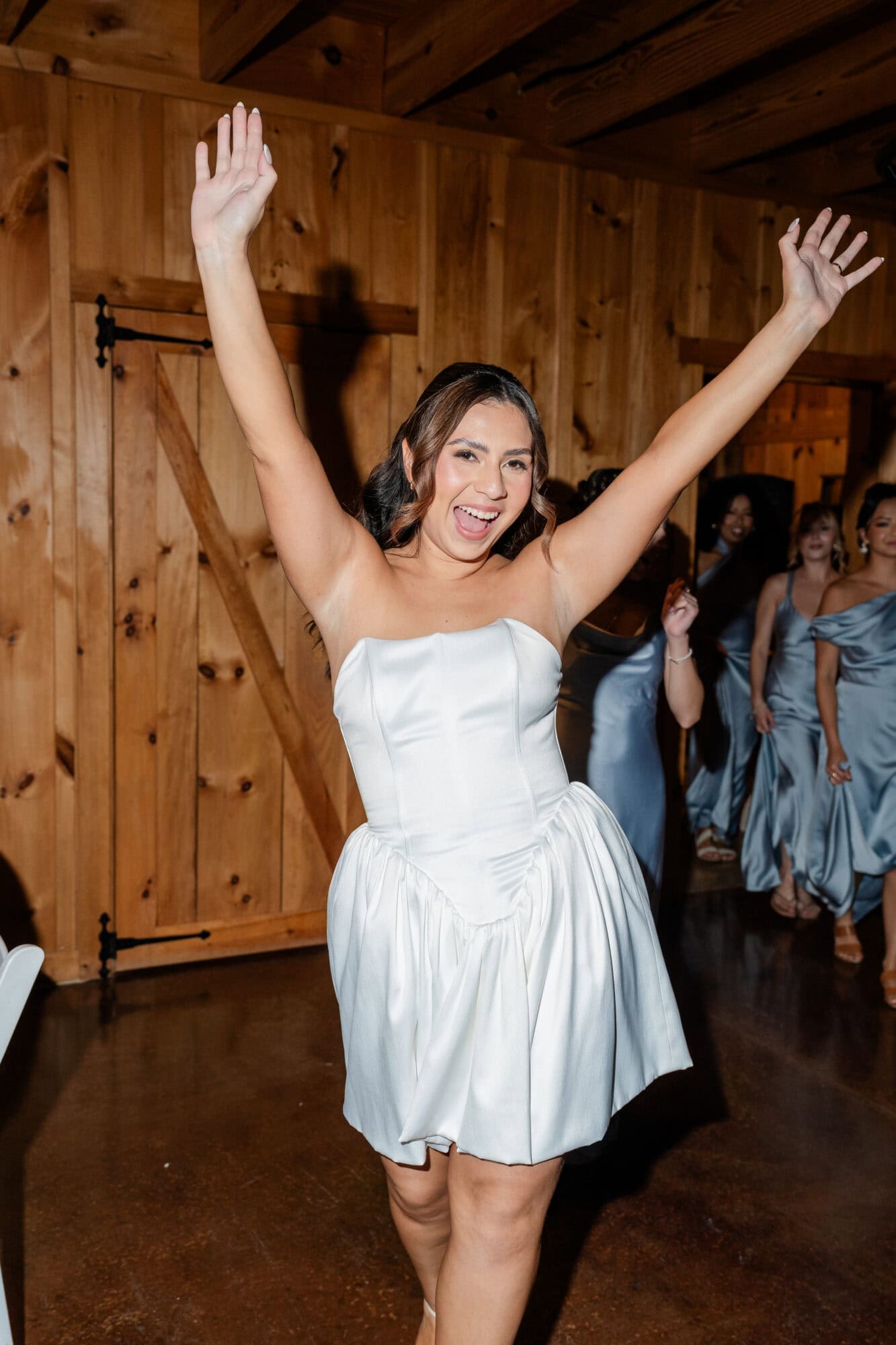 A woman in a strapless white dress smiles and raises her arms in celebration indoors, while other women in matching dresses stand behind her in a wooden room, perhaps gathering for a photo session inspired by a helpful Pricing Guide Template.