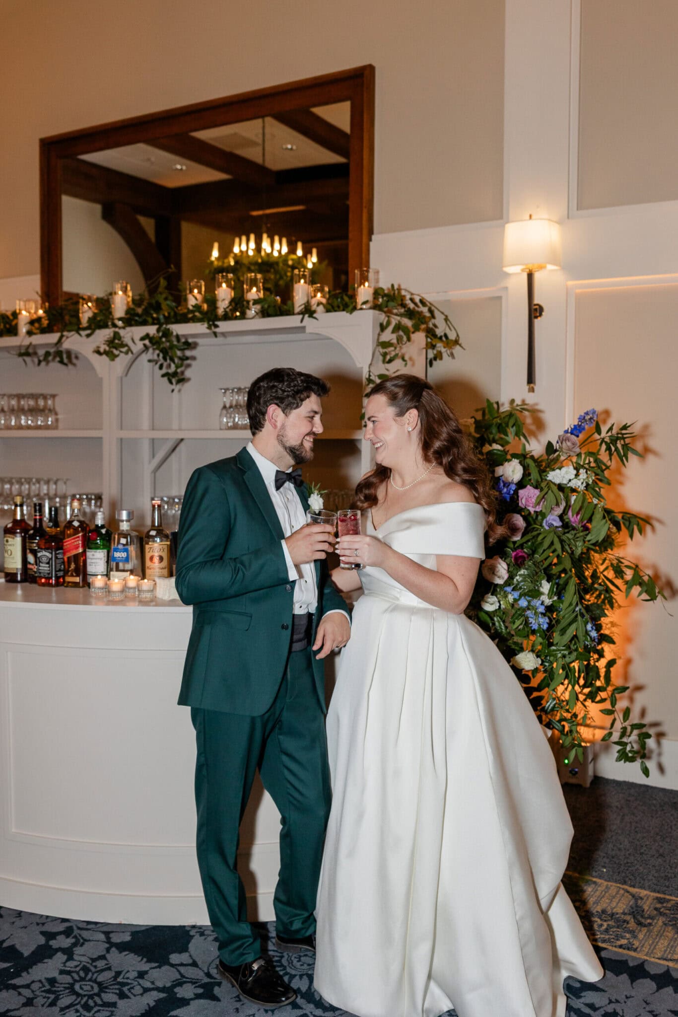 A bride in an off-the-shoulder white gown and a groom in a green suit smile at each other, holding drinks in front of a cozy wedding bar adorned with greenery and candles&mdash;capturing the warmth of cherished family moments.