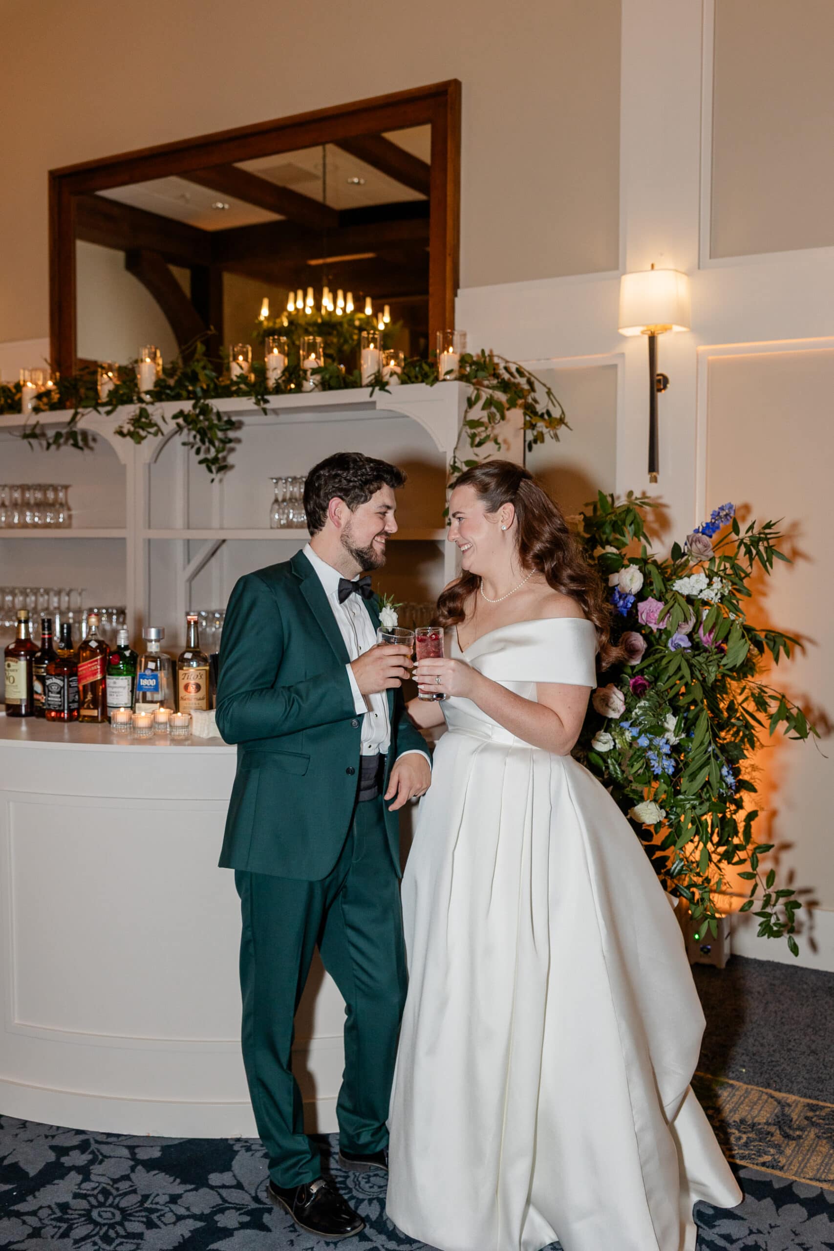 A bride in an off-the-shoulder white gown and a groom in a green suit smile at each other, holding drinks in front of a cozy wedding bar adorned with greenery and candles&mdash;capturing the warmth of cherished family moments.