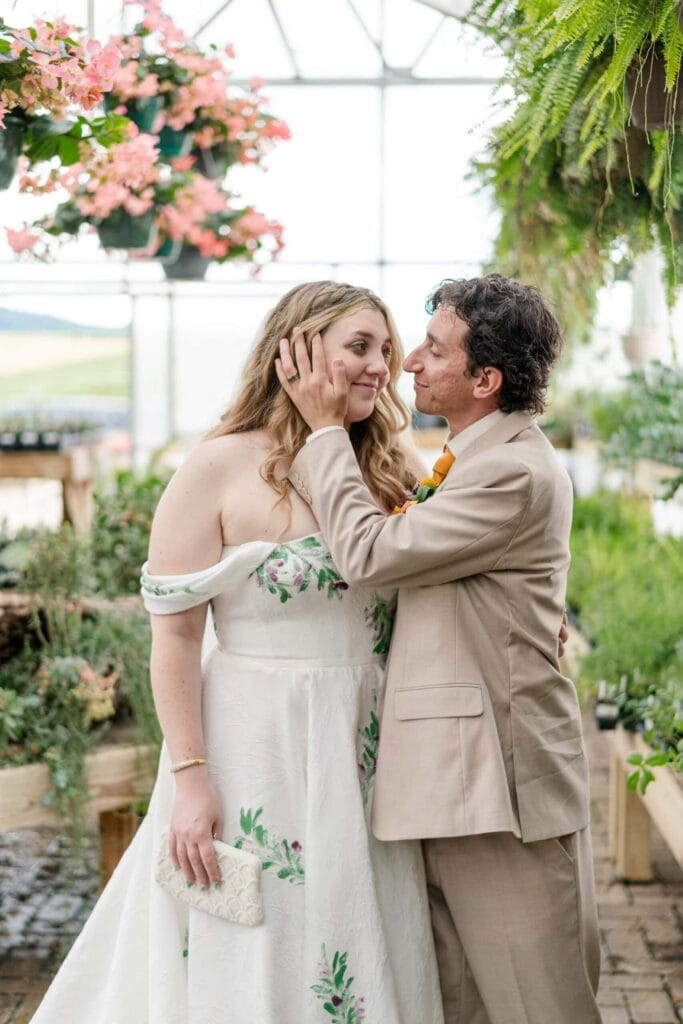 A man in a tan suit lovingly holds a woman&rsquo;s face and leans in to kiss her cheek. The woman, smiling in an off-the-shoulder white dress with vivid florals, stands with him in a greenhouse filled with lush plants and flowers.