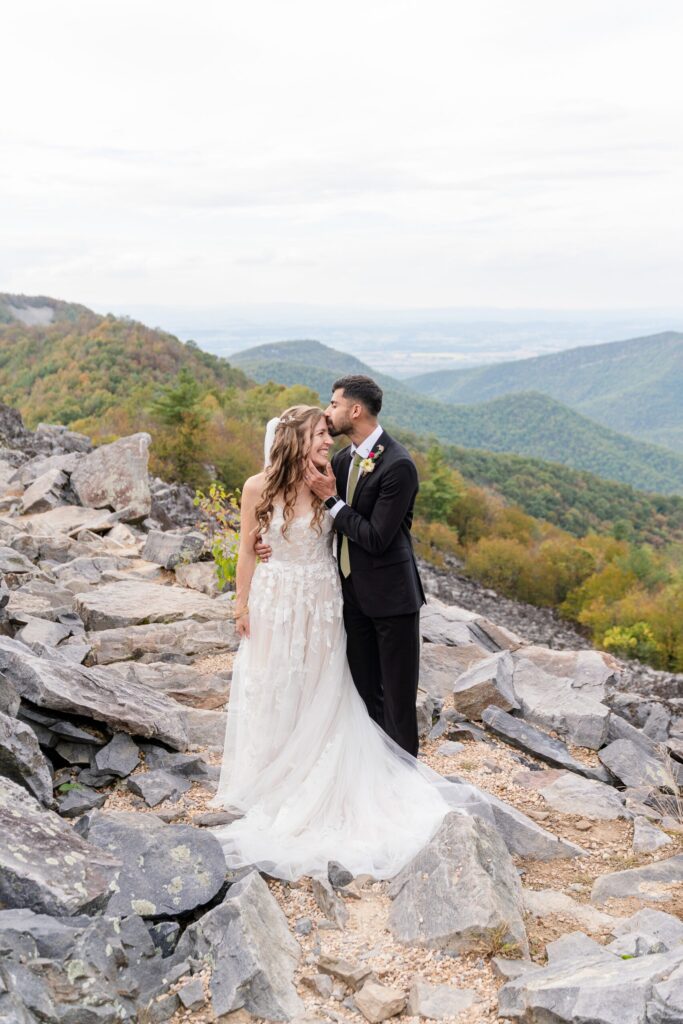 A bride and groom stand on rocky terrain at Raven’s Roost overlooking a scenic mountain landscape. The groom kisses the bride’s forehead as they smile, surrounded by greenery and distant hills—a perfect adventure elopement under a cloudy sky.