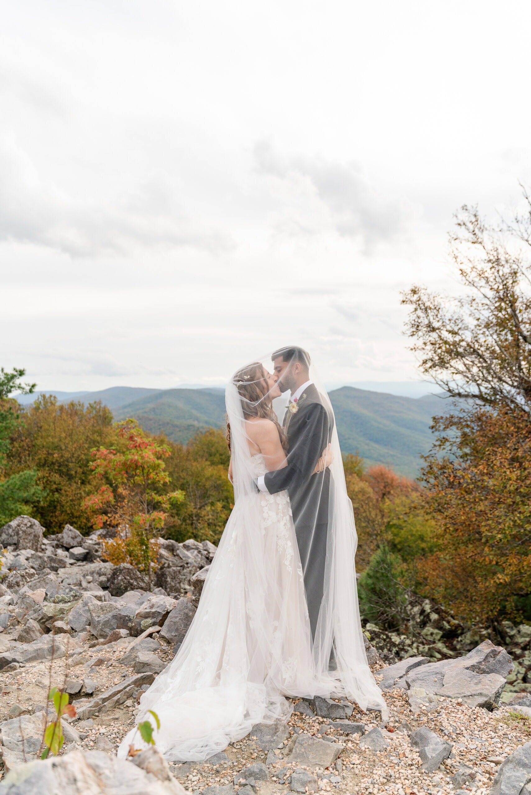 A bride and groom stand on rocky terrain at Raven&rsquo;s Roost, embracing and gazing at each other, surrounded by autumn trees and mountains in the background under a cloudy sky&mdash;a perfect adventure elopement setting.