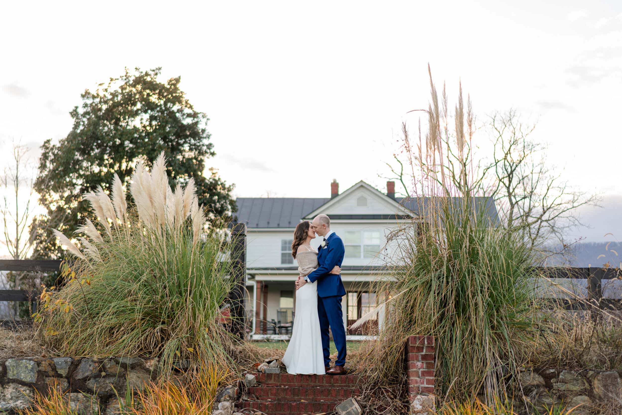 A bride and groom embrace and kiss on brick steps, surrounded by tall grasses and a white farmhouse—capturing the romantic charm of the Lewis Catherine House, a Virginia micro wedding venue, under a bright sky.