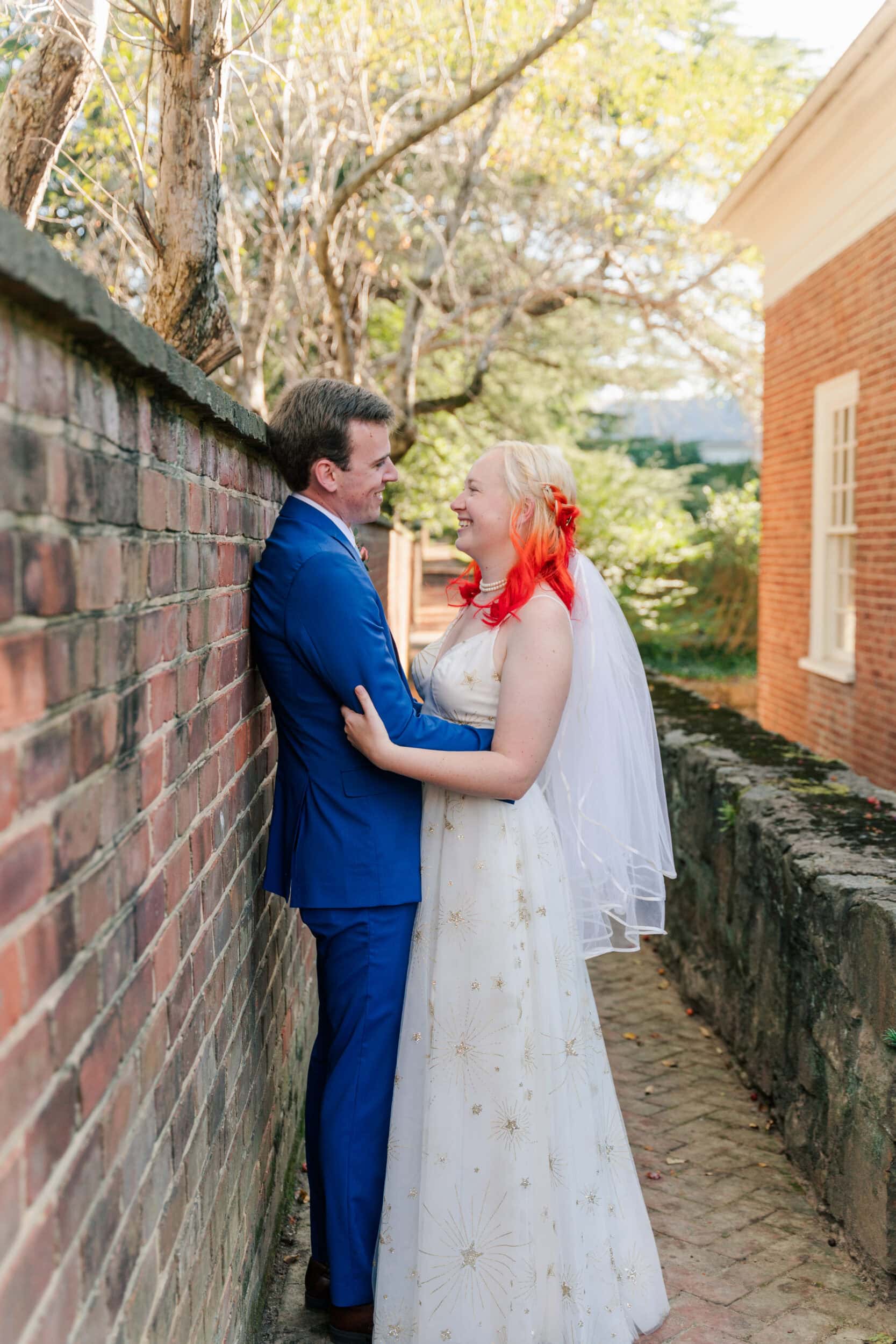 A bride in a white dress with orange hair and a veil smiles at her groom in a blue suit as they embrace beside a brick wall, enjoying their University of Virginia wedding outdoors on a sunny day.