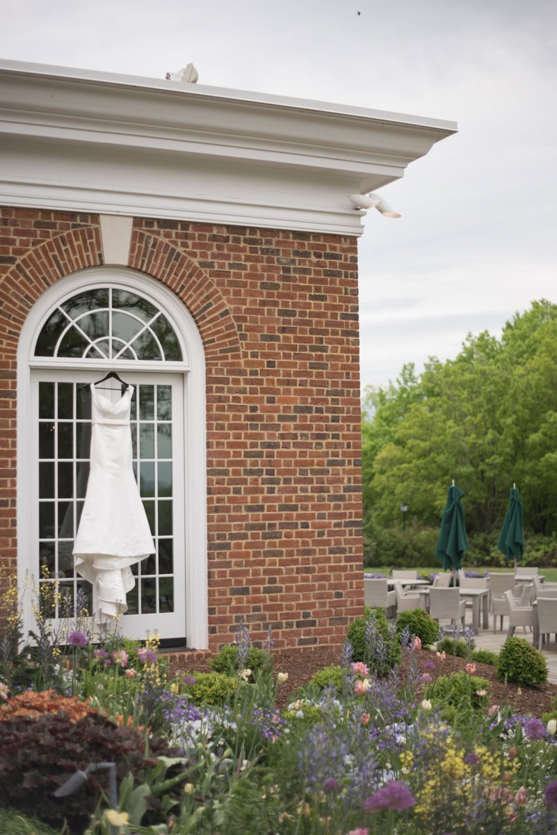 A white wedding dress hangs in front of a large arched window at Farmington Country Club, surrounded by a vibrant garden with colorful flowers. Two white doves perch on the building’s roof ledge—a perfect scene for anyone getting married.