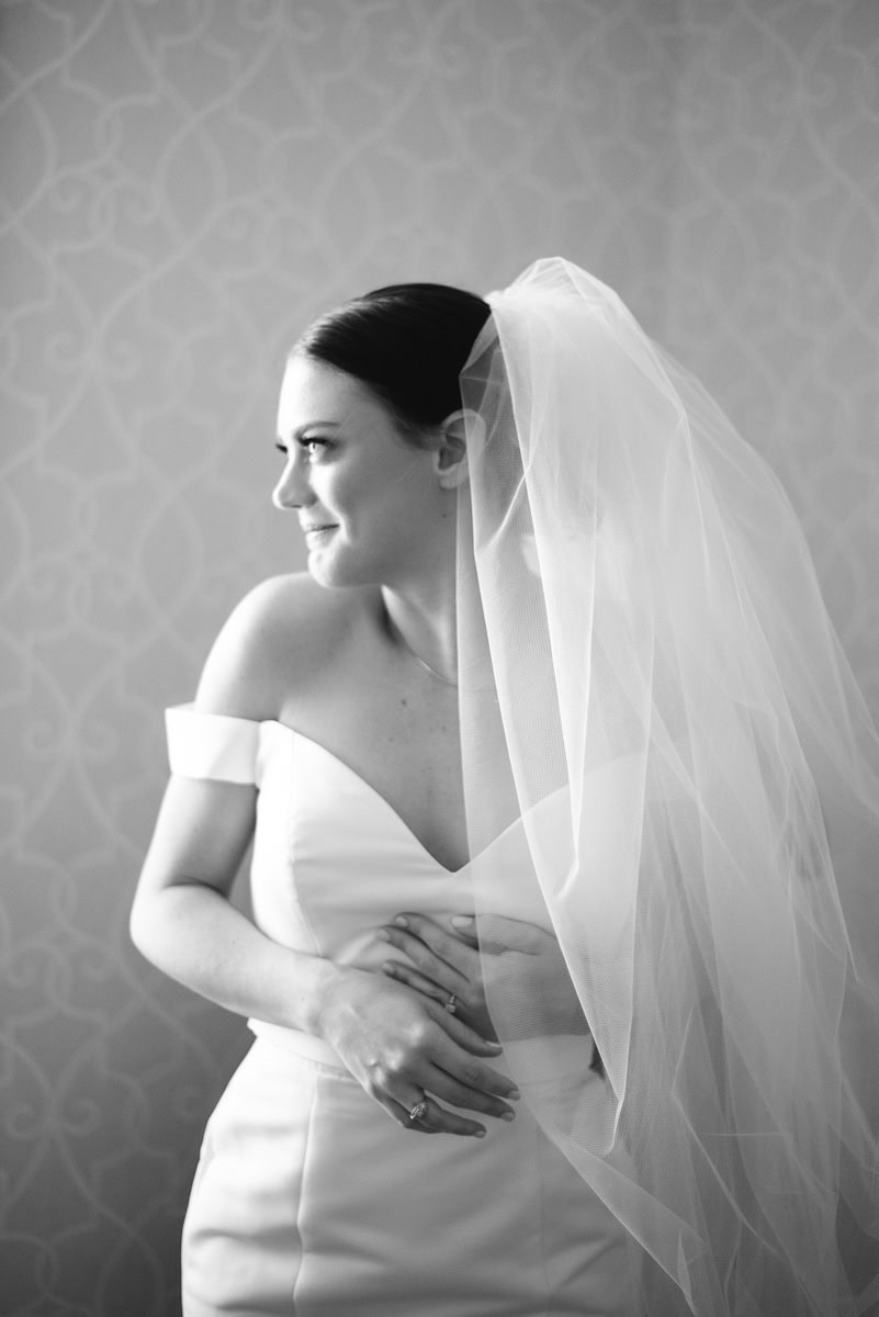 A bride in an off-the-shoulder wedding dress and veil smiles softly while looking to the side, her hands gently clasped at her waist. The black-and-white photo evokes timeless elegance—perfect inspiration for anyone getting married at Farmington Country Club.