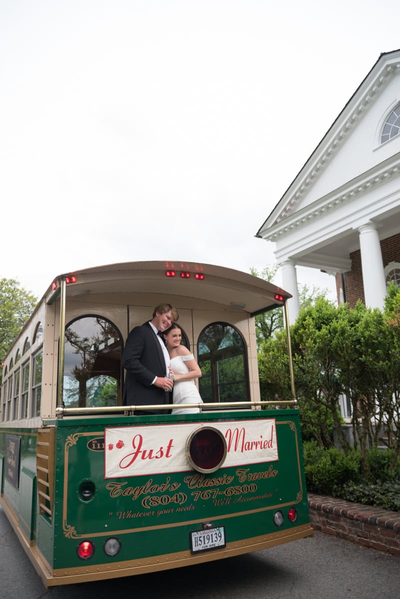 A newlywed couple stands on the back of a green trolley at Farmington Country Club, embracing and smiling beneath a "Just Married" sign. The trolley is parked near a white building and lush green trees—a perfect scene for any wedding guide.