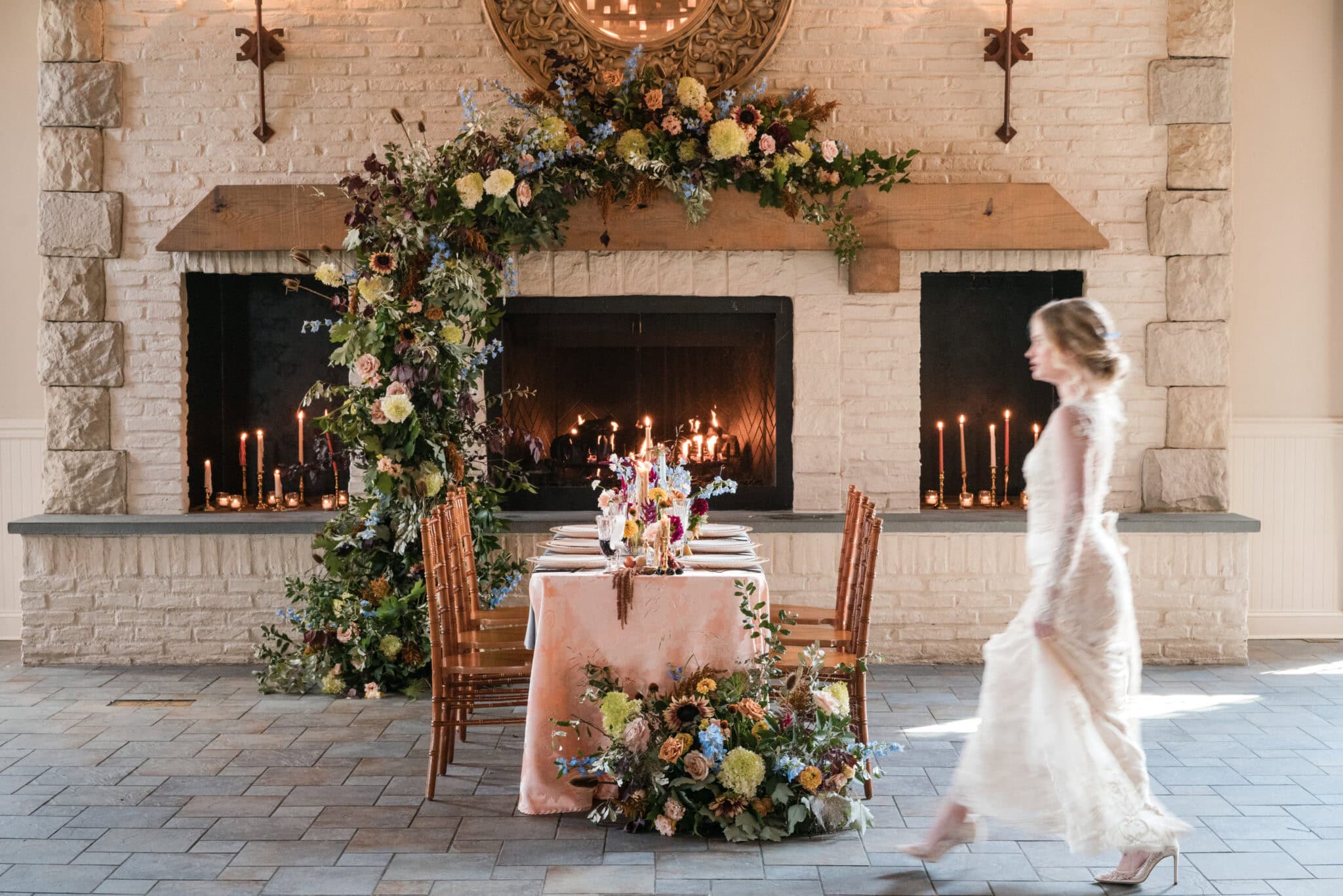 A blurred bride in a lace gown walks past an elegant wedding table for six, decorated with colorful flowers and candles, at Early Mountain Vineyards, with a fireplace adorned in lush blooms&mdash;perfect inspiration for your wedding guide.