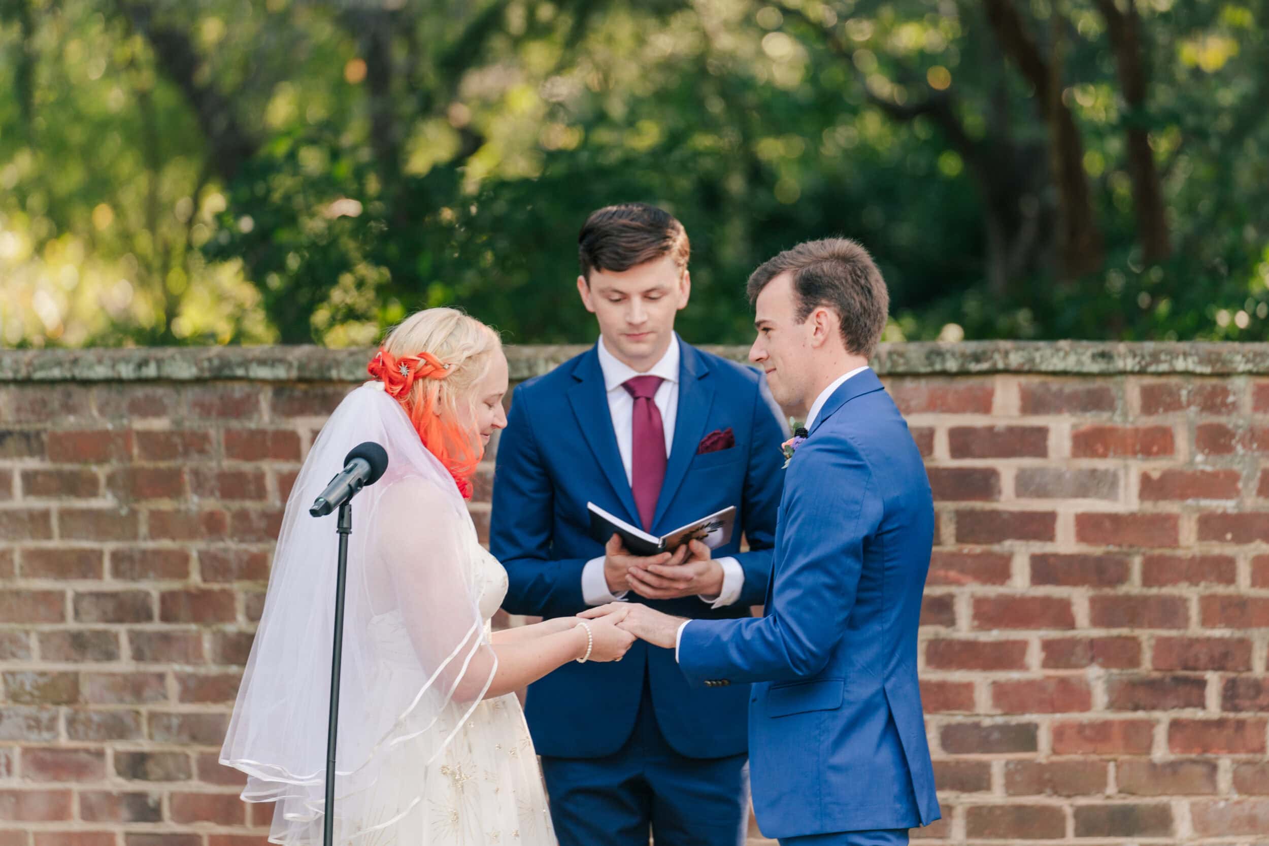 A bride and groom stand facing each other during an outdoor ceremony at the University of Virginia Gardens, exchanging rings as an officiant in a blue suit reads from a book. A brick wall and trees create a charming UVA wedding backdrop.