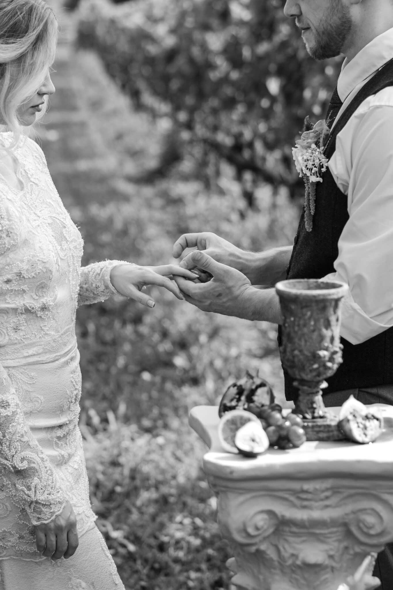 A bride and groom stand outdoors, getting married at Early Mountain as the groom places a ring on the bride’s finger. The bride wears a lace dress, with a decorative table of fruit and a goblet in the foreground. The scene is in black and white.
