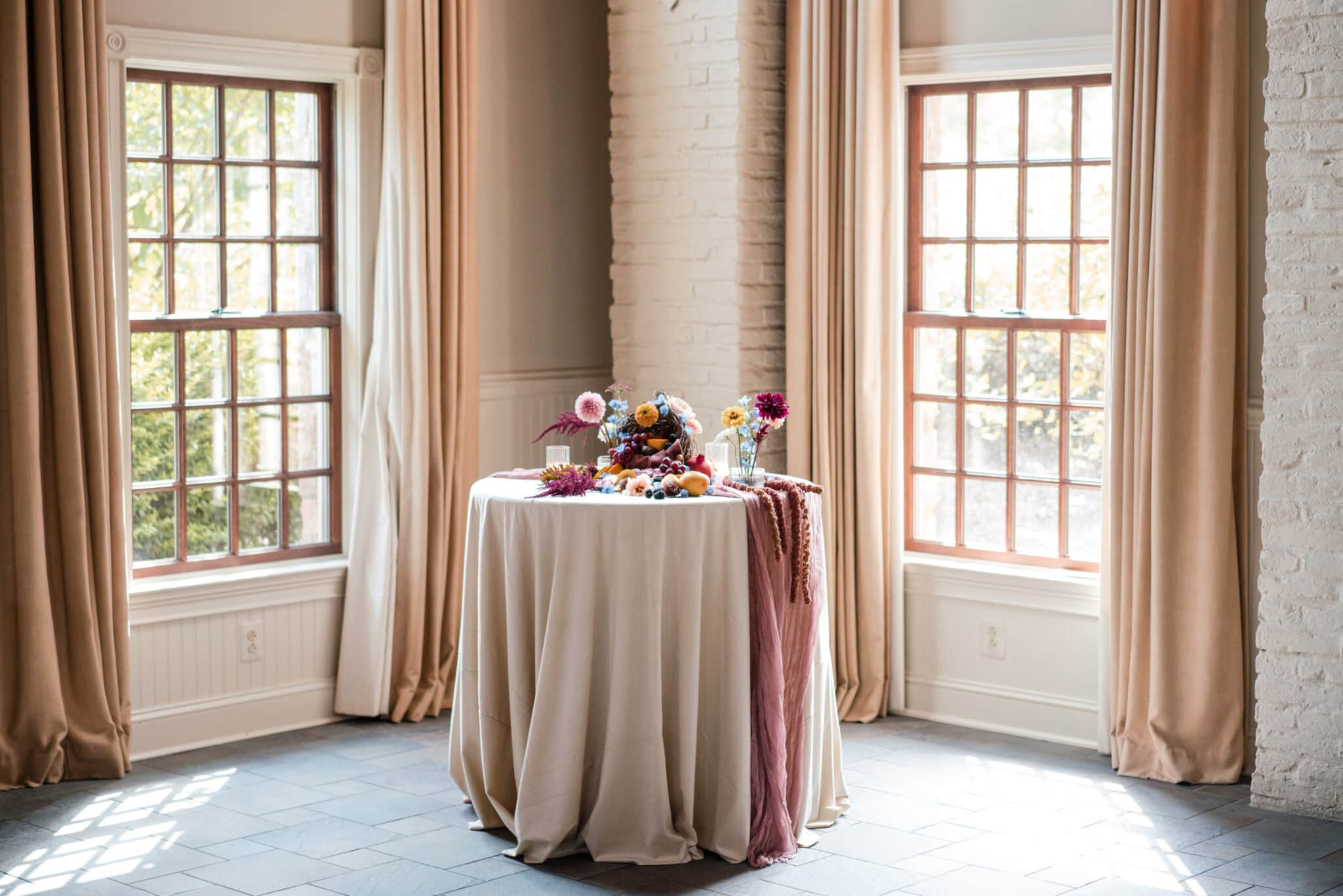 A round table with a cream tablecloth stands between two large windows with beige curtains, decorated for getting married with flowers and small colorful items, in a bright, elegant room at Early Mountain Vineyards.