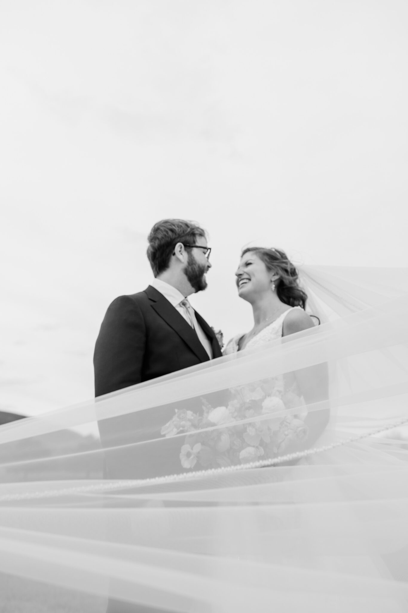 A bride and groom smile at each other outdoors in Lexington, surrounded by flowing veil fabric. The image is in black and white, giving it a soft, romantic feel as the bride holds a bouquet of colorful florals.