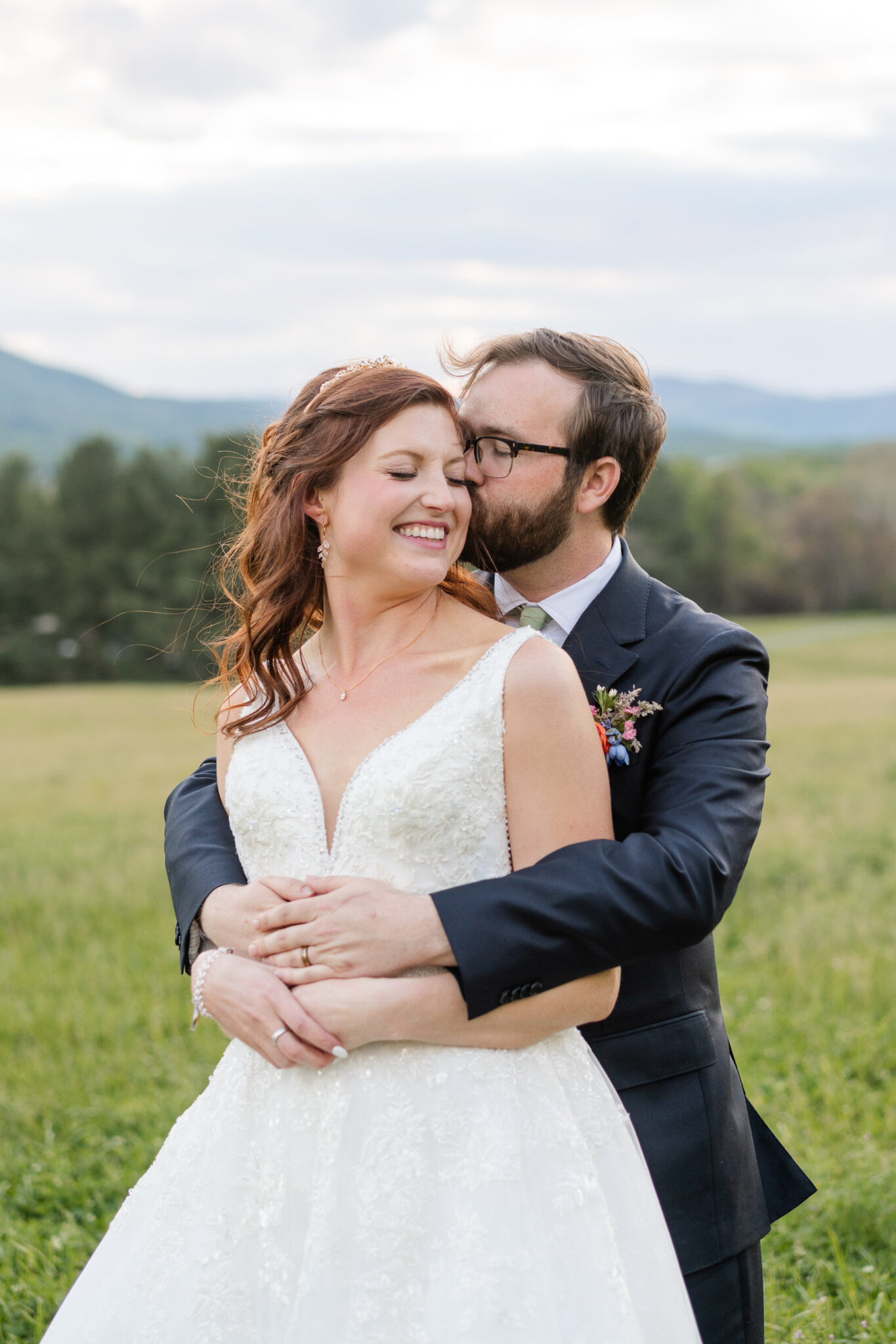 A groom in a suit embraces and kisses a smiling bride in a white wedding dress outdoors, surrounded by colorful florals at Big Spring Farm, with green grass, trees, and mountains in the background.