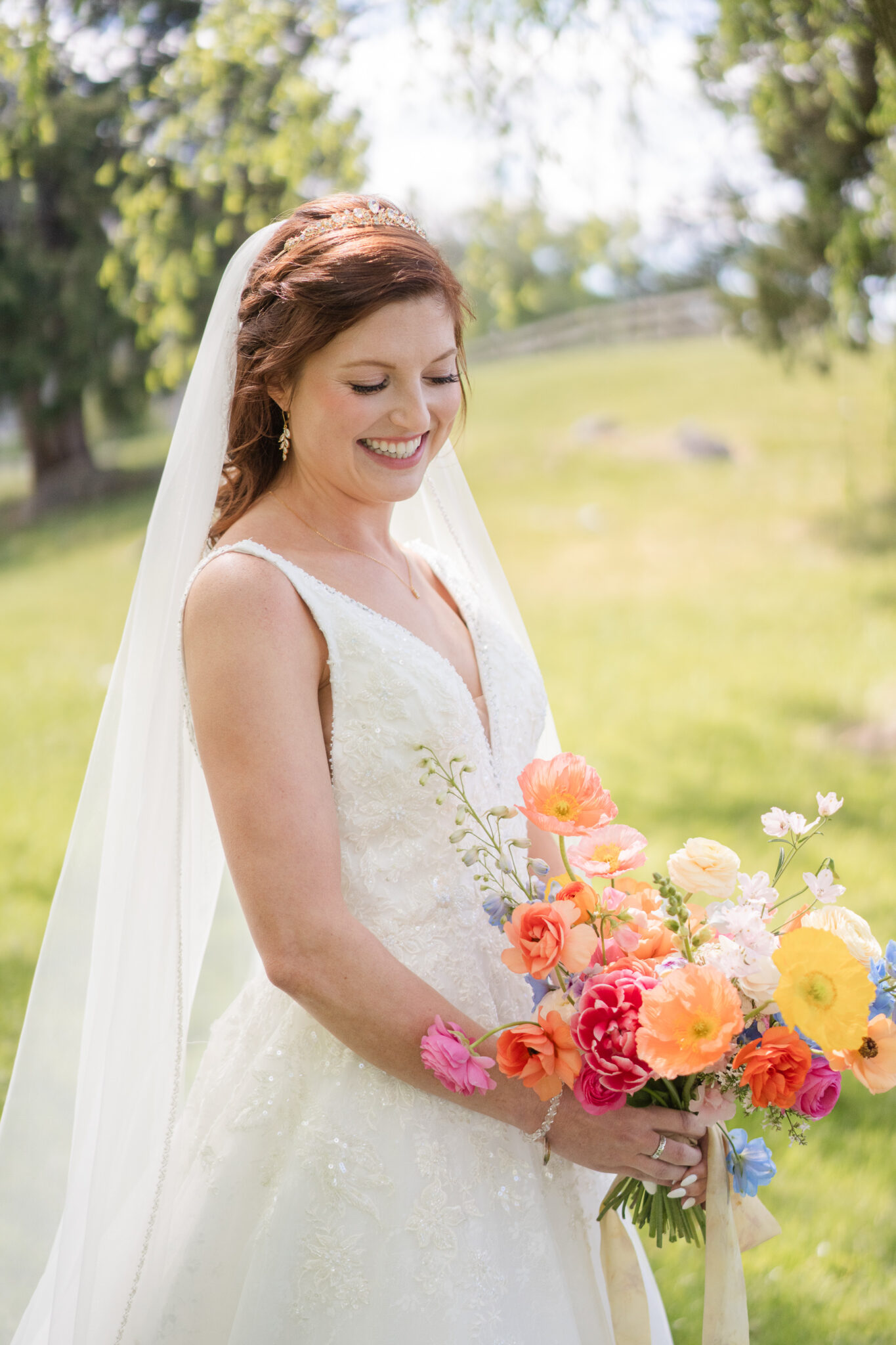 A bride in a white lace gown and veil smiles while holding colorful florals outdoors on a sunny day at Big Spring Farm in Lexington, with green trees and grass in the background.