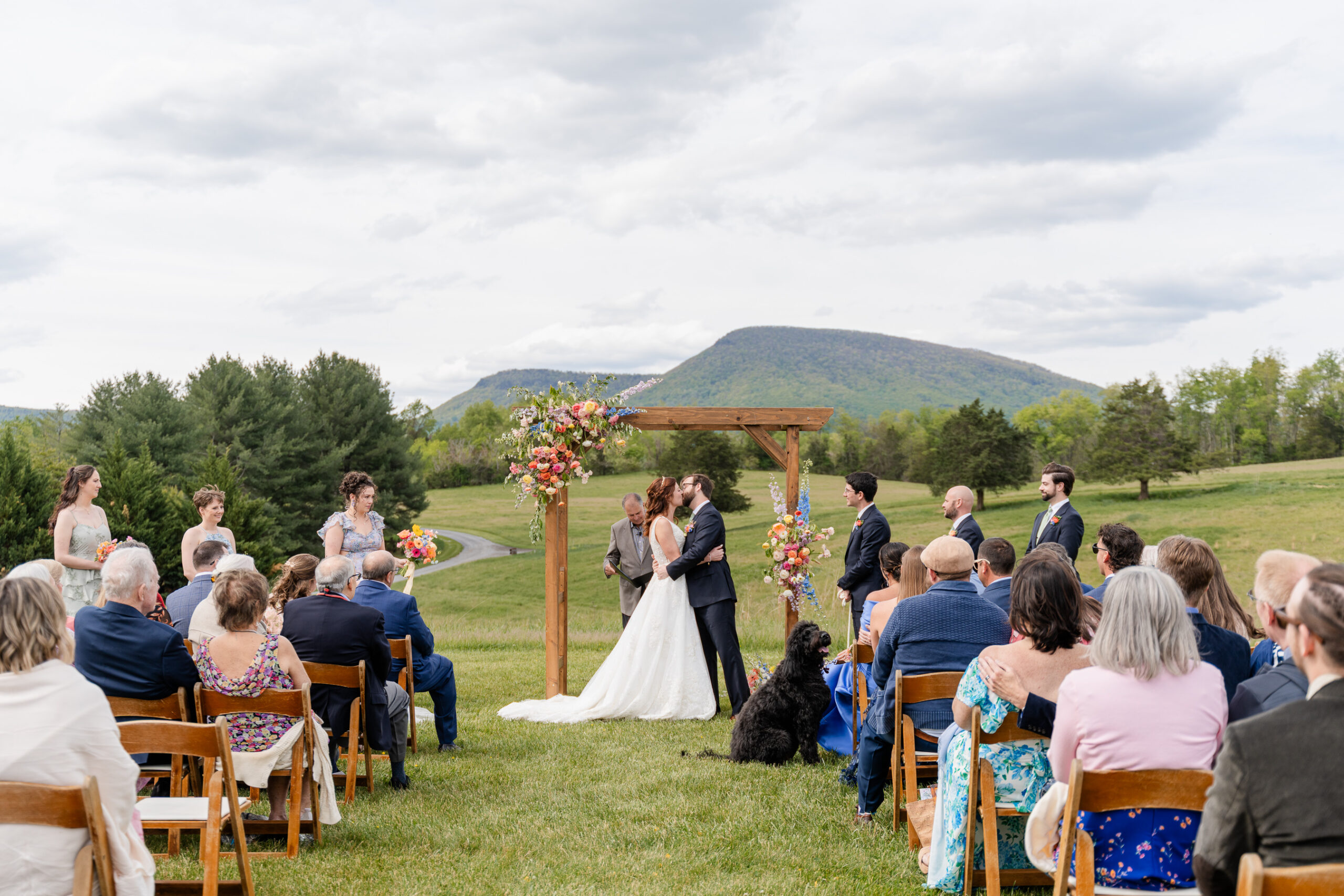 A couple kisses under a wooden arch decorated with colorful florals during an outdoor wedding ceremony at Big Spring Farm in Lexington, with guests seated on wooden chairs and mountains visible in the background.