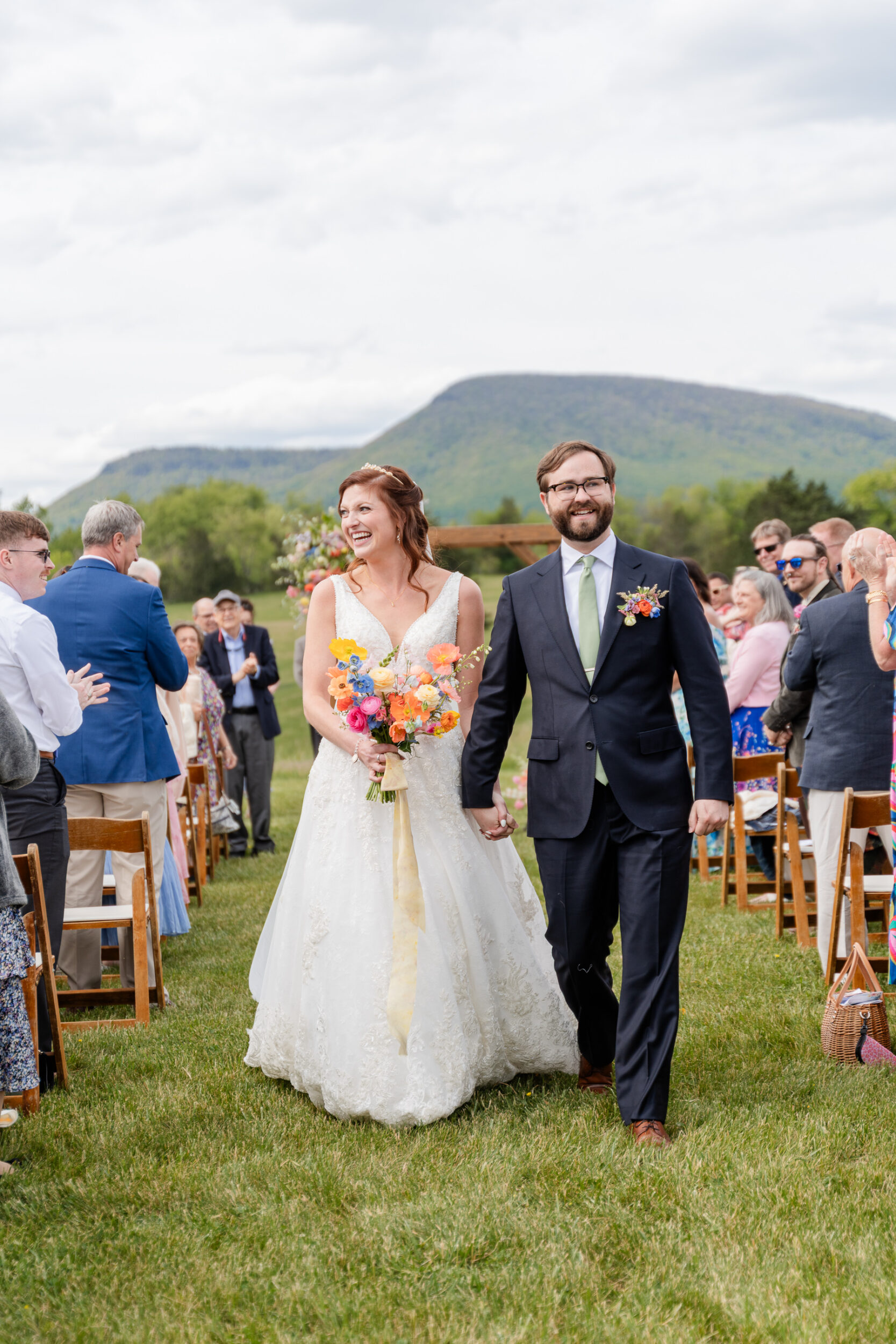 A bride and groom walk hand in hand down an outdoor aisle at Big Spring Farm, smiling joyfully. Guests sit on wooden chairs, clapping. The bride carries colorful florals, with green mountains visible in the background under a cloudy sky.