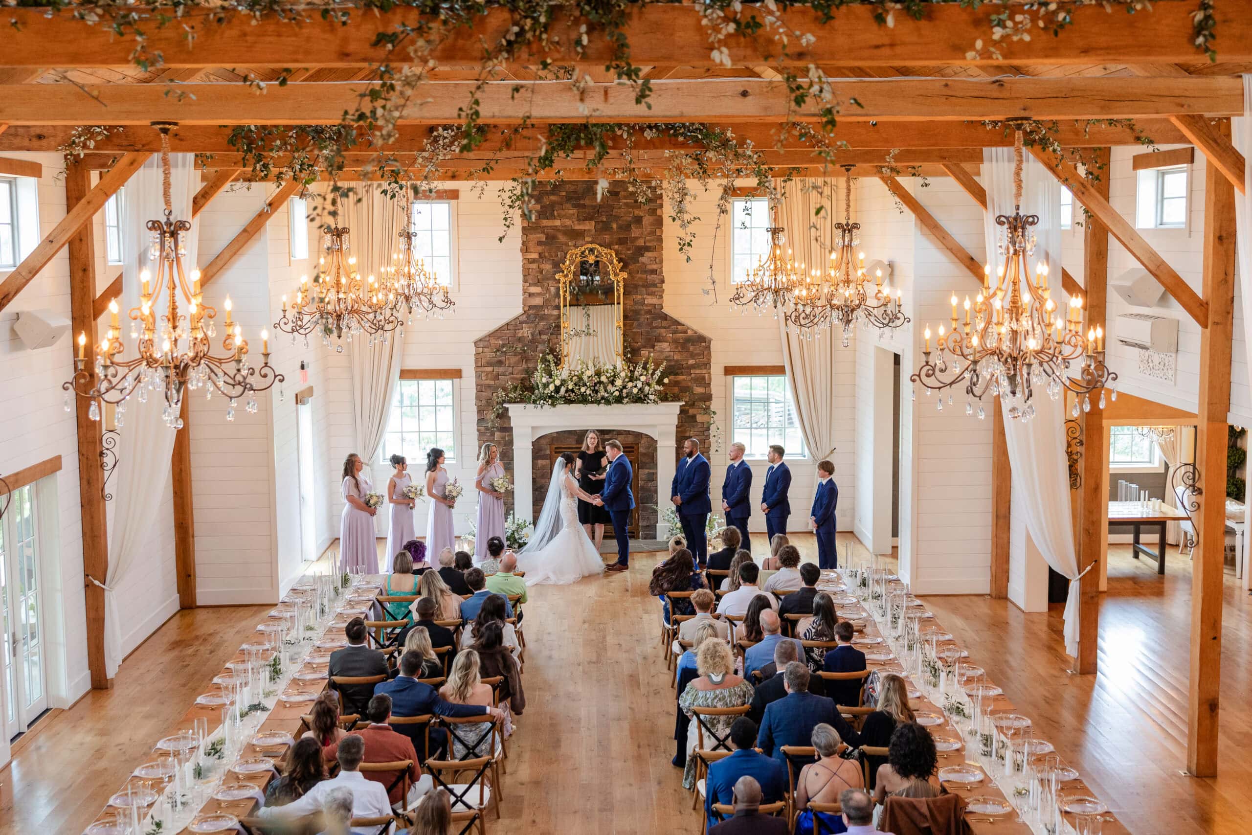 A bride and groom stand at the altar in a bright, rustic wedding venue at The Secusion in Lexington, with chandeliers, floral garlands, and guests on both sides. Bridesmaids in lavender and groomsmen in blue look on during this rainy wedding day.