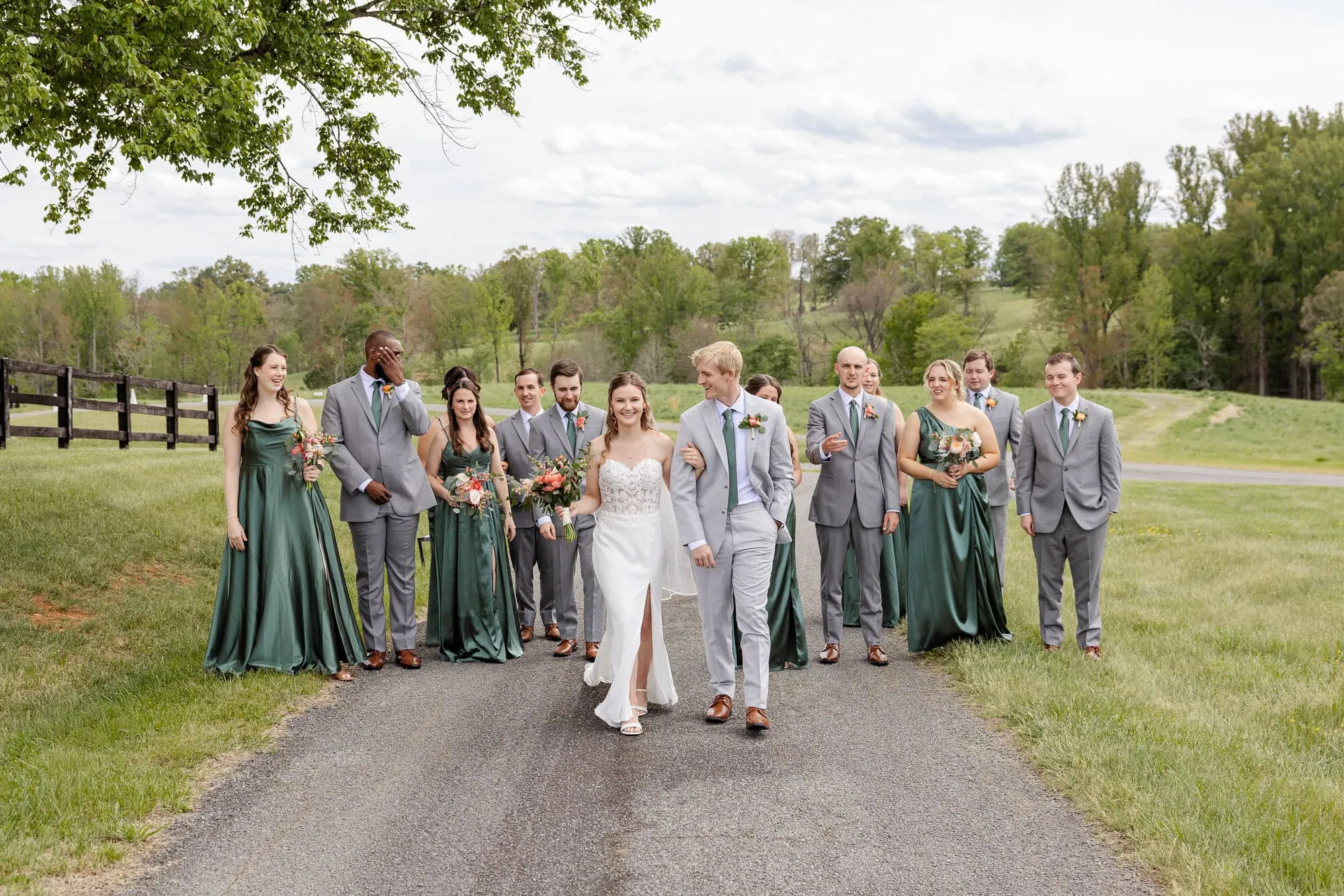 A bride and groom walk hand in hand down a paved path at their Tiffin Barn wedding, smiling, surrounded by their wedding party in green dresses and gray suits, with trees and grassy fields in the background.