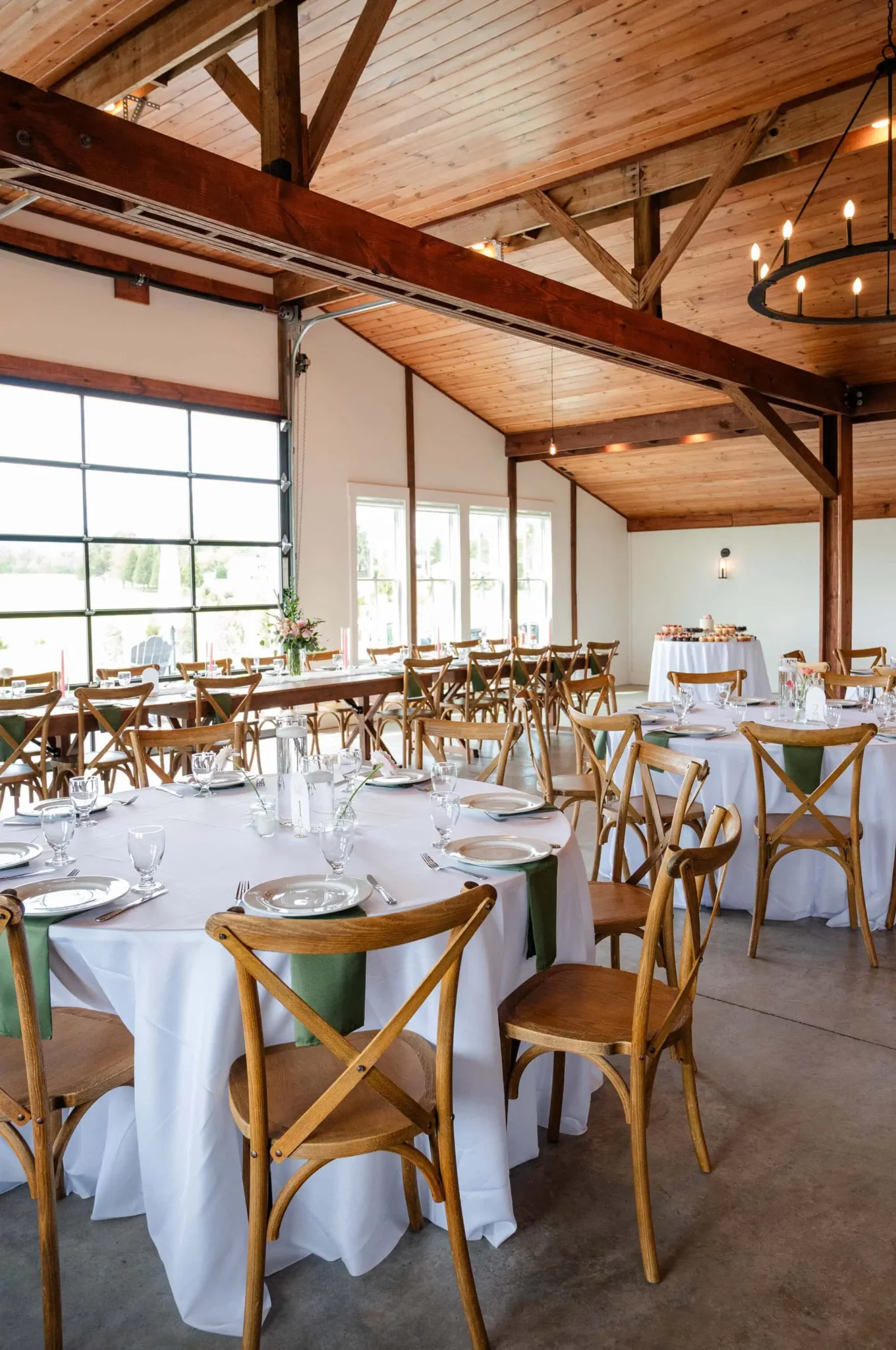 A bright event space at Tiffin Barn, with wooden beams, round tables covered in white tablecloths, wooden chairs with green napkins, set for a meal, and large windows letting in natural light—perfect for unforgettable Tiffin Barn wedding photos.